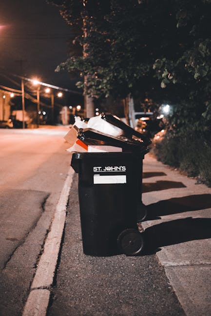 An outdoor scene showing a large, rectangular red metal waste container with visible signs of rust and chipped paint, positioned against a dark green wall and adjacent to a light grey concrete wall. T